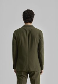 Chaqueta de traje verde olivo vista desde la parte de atrás, con una abertura sencilla, solapas con muesca y costuras sutiles en los hombros y las mangas.