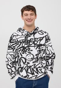 Hoodie bianco e nero con una stampa in stile graffiti contenente vari elementi testuali e una faccina sorridente, realizzato in tessuto con una vestibilità standard.