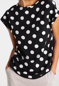 Svart kortärmad topp med vita prickar, med en bröstficka och en lös passform, tillverkad av ett mjukt tyg.