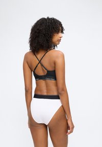 Kvinde med krøllet hår iført en sort Calvin Klein-bh med krydset strop og hvide trusser med høj talje, set bagfra mod en ensfarvet baggrund.