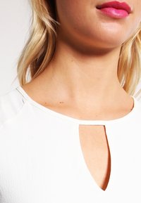 Weißes Oberteil mit rundem Ausschnitt und zentralem Schlüsselloch-Ausschnitt. Glatte Textur und minimalistisches Design. Hintergrund unscharf.