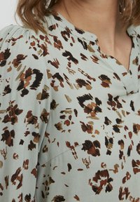 Blouse vert clair avec col en V et manches bouffantes, présentant un motif léopard marron et noir sur un tissu doux.