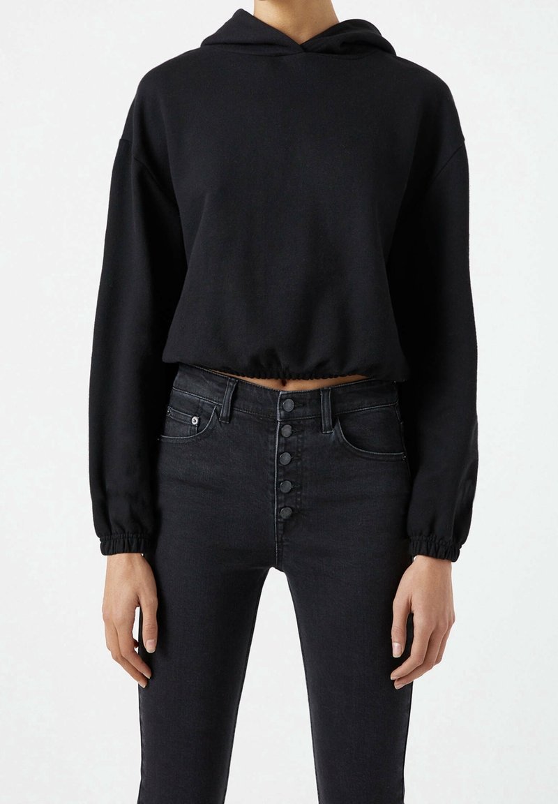 Personne portant un sweat à capuche court noir avec ourlet élastique et un jean noir taille haute avec plusieurs boutons devant sur un fond uni.