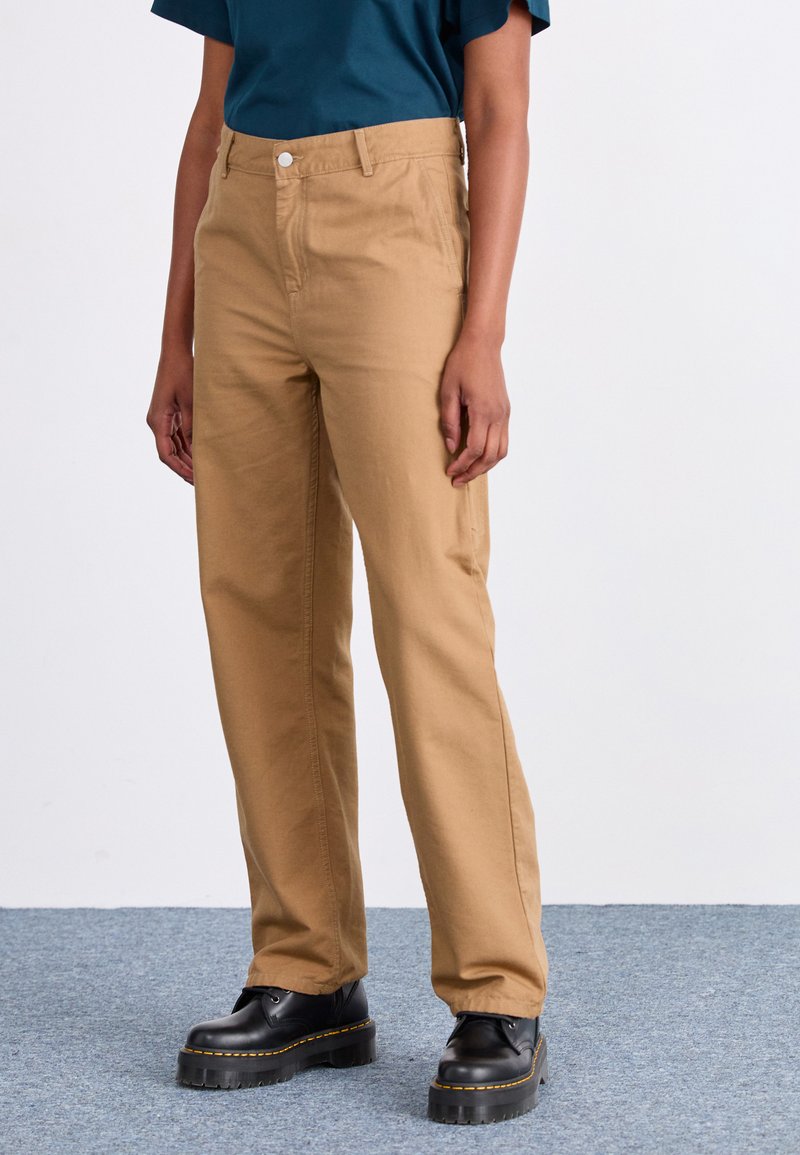 Khaki cotton trousers with straight legs, a front button closure, and side pockets. Worn with black platform boots.