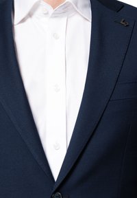 Granatowa marynarka z wyłogami, z białą koszulą z guzikami pod spodem. Tkanina ma gładką fakturę i elegancki krój.