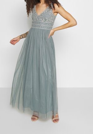 Femme portant une robe sans manches avec un corsage gris orné et une jupe fluide, debout avec une main sur la hanche, accompagnée de sandales ouvertes beiges.