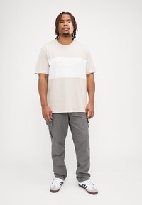 Beige katoenen t-shirt met witte horizontale streep en reliëftekst, gecombineerd met grijze cargobroek en witte sneakers met zwarte accenten.