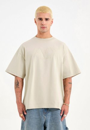 Hombre con cabello rubio corto, vistiendo una camiseta beige grande de AVX y jeans azules, mostrando tatuajes en ambos antebrazos contra un fondo blanco.