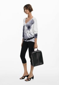Schwarze Tragetasche mit kontrastierenden Nähten, die ein strukturiertes Design und Obergriffe aufweist. Kombiniert mit einem weißen und grauen Grafik-Langarmoberteil und Leggings.