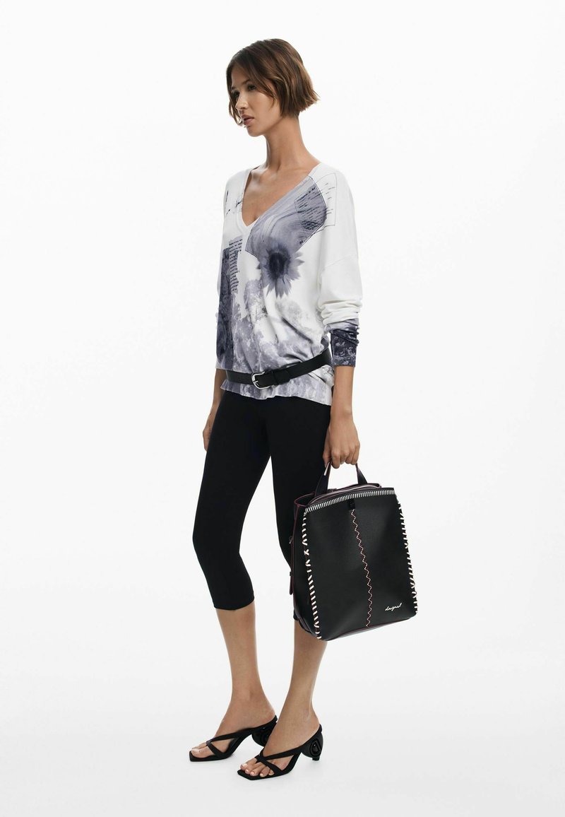 Schwarze Tragetasche mit kontrastierenden Nähten, die ein strukturiertes Design und Obergriffe aufweist. Kombiniert mit einem weißen und grauen Grafik-Langarmoberteil und Leggings.