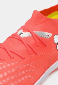 Lyserød fodboldstøvle med tekstureret overflade, tætsidende pasform og sølvfarvede dekorative detaljer. Snørebånd er tydeligt fremhævet mod den levende baggrund.