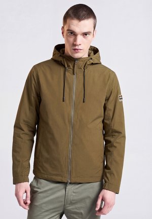 Mann trägt eine olivgrüne Kapuzenjacke mit Frontreißverschluss und Kordelzügen, steht vor einem einfarbigen, hellen Hintergrund.