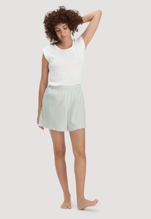 Frau mit lockigem Haar, die ein weißes ärmelloses Oberteil und hellgrüne gestreifte Shorts trägt, steht barfuß da und hat einen Arm hinter dem Kopf erhoben.