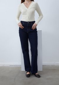 Cremfarbener Langarmoberteil mit V-Ausschnitt und gedrehter Vorderseite, kombiniert mit hochtaillierten, dunkelblauen Jeans im Bootcut-Stil und braunen flachen Schuhen.