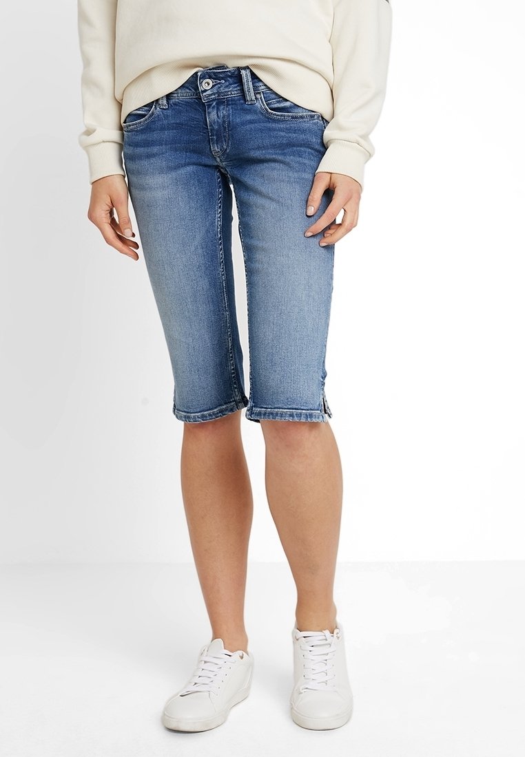 Person klädd i knälånga blå denimshorts, vita sneakers och en beige långärmad topp, stående mot en vit bakgrund.