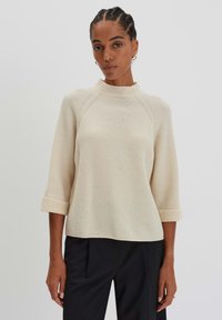 Maglione a coste color crema con un collo alto, maniche a tre quarti e polsini arrotolati, abbinato a pantaloni scuri.