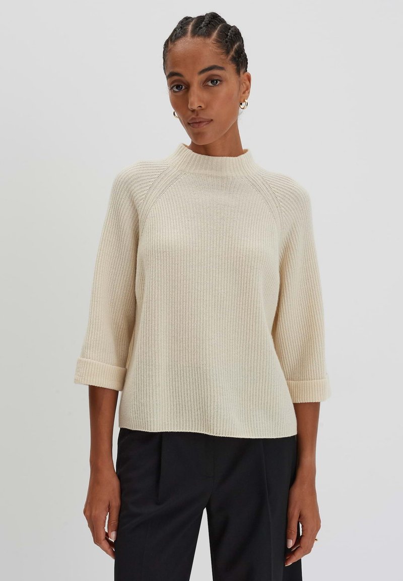 Maglione a coste color crema con un collo alto, maniche a tre quarti e polsini arrotolati, abbinato a pantaloni scuri.