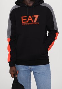 Sudadera con capucha negra, con paneles en los hombros grises y detalles en naranja. Presenta un logo bordado de "EA7 Emporio Armani" en el frente.