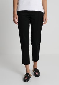 Pantalones negros, ajustados y de corte corto. Tela suave con costuras delanteras y sin bolsillos visibles. Combinados con mocasines negros.