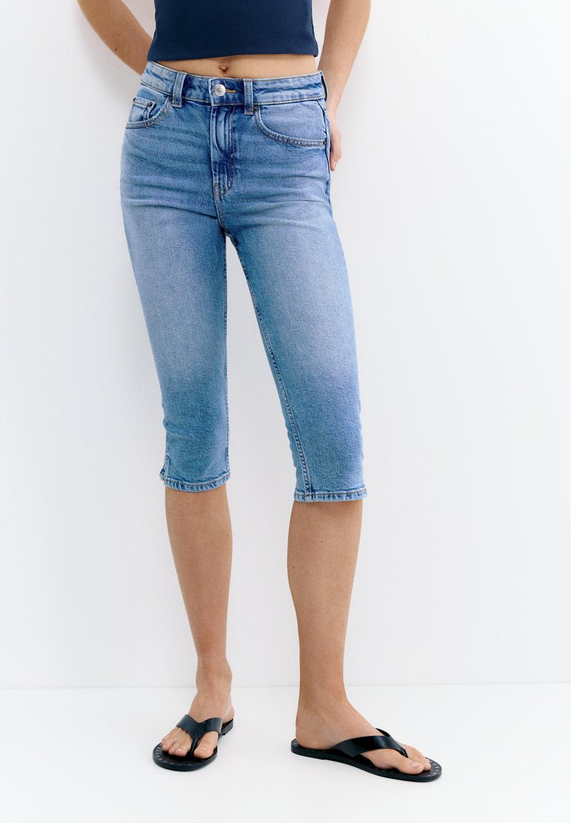 Persoon draagt mid-rise, lichtgewassen denim capri jeans en zwarte slip-on sandalen, staand tegen een eenvoudige witte achtergrond.