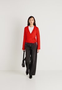 Červený zabalovací kardigan s dlouhými rukávy, texturovaný úplet. Kombinovaný s černými džínami s vysokým pasem a černou kabelkou, nošený přes bílou košili.