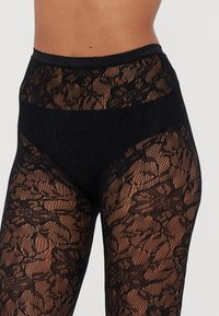 Leggings en dentelle noire avec un motif floral, design taille haute, texture semi-transparente et une ceinture noire solide.