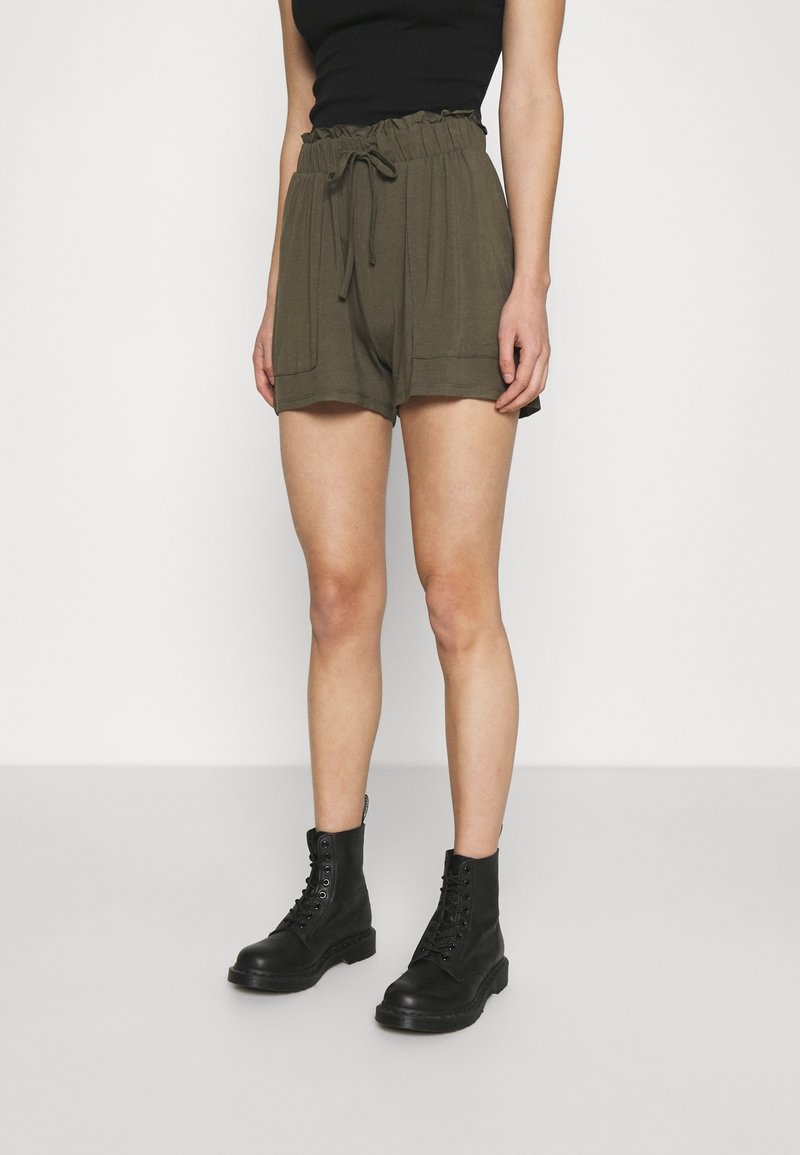 Femme portant un short taille haute vert olive avec poches avant et des bottes noires à lacets, debout sur un sol gris clair.
