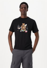 Zwarte T-shirt met korte mouwen, voorzien van een berenafbeelding te midden van campingelementen in levendige kleuren, tegen een achtergrond van een bos.