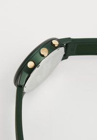 Grön klocka med rund klocka, guldfärgade detaljer och strukturerad vit urtavla. Slät silikonrem för komfort och hållbarhet.