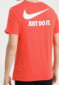 Červená košile s krátkým rukávem a bílým logem Nike s textem "JUST DO IT." na zádech, kterou nosí osoba otočená zády.