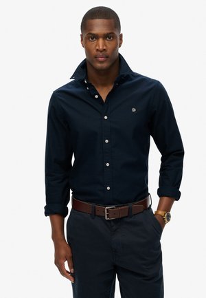 Navy button-up shirt with a collar, long sleeves rolled at the cuffs, and a small embroidered logo. Paired with dark pants and a brown belt.