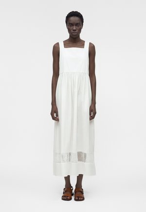 Γυναίκα που φοράει λευκό αμάνικο μίντι φόρεμα με τετράγωνη λαιμόκοψη και λεπτομέρεια πλεξίματος κοντά στο τελείωμα, φοράει καφέ φλατ σανδάλια.