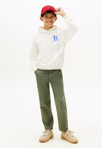 Felpa bianca con cappuccio e logo blu, pantaloni verde oliva e sneakers beige. Cappellino rosso con un piccolo logo sulla parte anteriore.