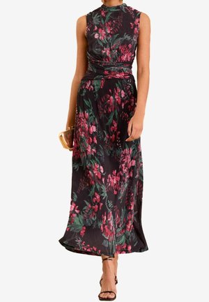 Kobieta w bezrękawowej, ciemnej sukience midi w kwiaty z marszczonym pasem, trzyma złotą kopertówkę i nosi czarne sandały na obcasie z paskami.