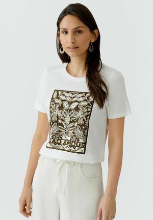 Vrouw draagt een wit T-shirt met een bruin en beige botanisch en vogelmotief en witte broek met trekkoord, staand tegen een lichte achtergrond.