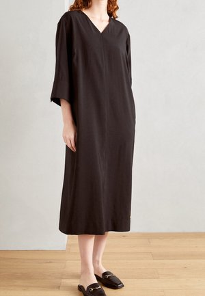 Frau trägt ein lockeres, schwarzes Midikleid mit dreiviertel Ärmeln und schwarze Slipper mit goldenen Verzierungen, steht auf einem Holzboden.