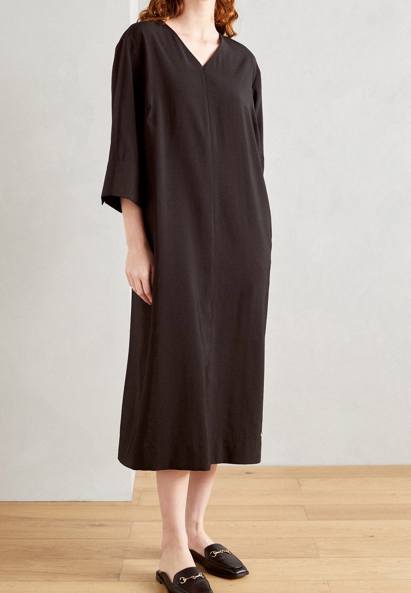 Femme portant une robe midi noire ample à manches trois-quarts et des mocassins noirs à enfiler avec des détails dorés, debout sur un sol en bois.
