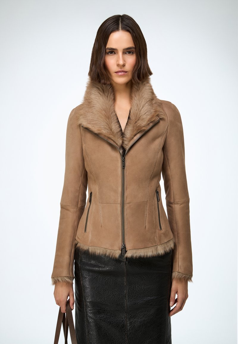Veste en daim beige avec col en fourrure, coupe ajustée, fermeture éclair frontale et poches latérales zippées, associée à une jupe texturée noire.
