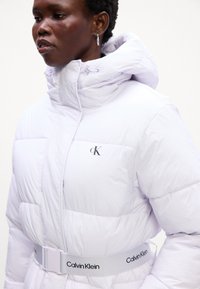 Biely nafúkaný kabát s vysokým golierom, prešívaným dizajnom a logom Calvin Klein. Obsahuje sťahovaný pás s označeným opaskom. Mäkká textura.