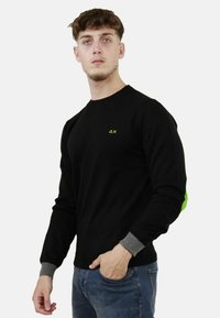 Felpa nera a maniche lunghe con un logo verde sul petto e polsini grigi. Tessuto morbido e design aderente. Il modello indossa jeans blu.