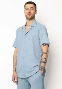 Chemise à manches courtes bleu clair en tissu texturé, avec col et boutons sur le devant, assortie à un pantalon coordonné.