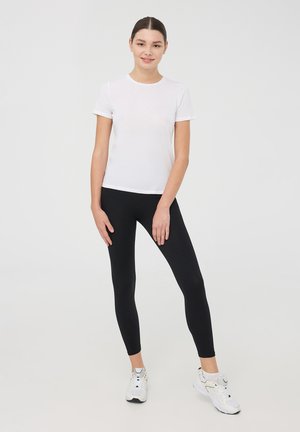 Biała koszulka z krótkim rękawem w połączeniu z czarnymi legginsami, charakteryzująca się dopasowanym krojem. Modelka ma na sobie białe buty sportowe. Proste, jednolite kolory.