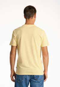 Hellgelbes Baumwoll-T-Shirt mit kurzen Ärmeln, rundem Ausschnitt und einem kleinen Logotag an der Seite; getragen mit blauen Denim-Jeans.