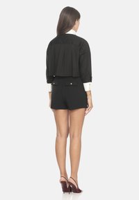 Cropped shirt nera con polsini bianchi, abbinato a pantaloni corti neri a vita alta con due tasche posteriori. Il modello indossa tacchi bordeaux.
