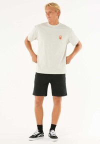 Lys grå t-shirt med korte ærmer og et lille rødt logo på venstre bryst, parret med sorte denimshorts og sorte sneakers med hvide detaljer.