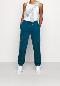 Pantalones de chándal verde azulado con puños elásticos, que cuentan con parches en contraste y un eslogan impreso. Combinados con una camiseta sin mangas blanca y un cárdigan gris.