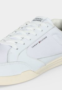 Baskets basses blanches avec lacets blancs, bout perforé, semelle beige, et marquage « Tommy Hilfiger » sur la languette et les côtés.