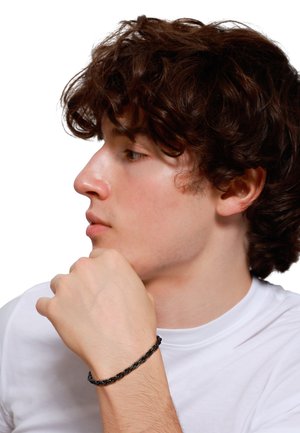Schwarzes geflochtenes Armband an einem Handgelenk, mit einem gedrehten Design. Die Person hat zerzaustes braunes Haar und trägt ein weißes Hemd.