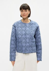 Μπουφάν με επένδυση σε μπλε χρώμα με γεωμετρικό σχέδιο, στρογγυλή λαιμόκοψη και μπροστινό φερμουάρ. Μακρυά μανίκια με χαλαρή εφαρμογή.