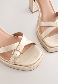 Gold metallic platform sandals with a square toe, crisscross straps, and a chunky heel. Features a decorative buckle on the strap.
